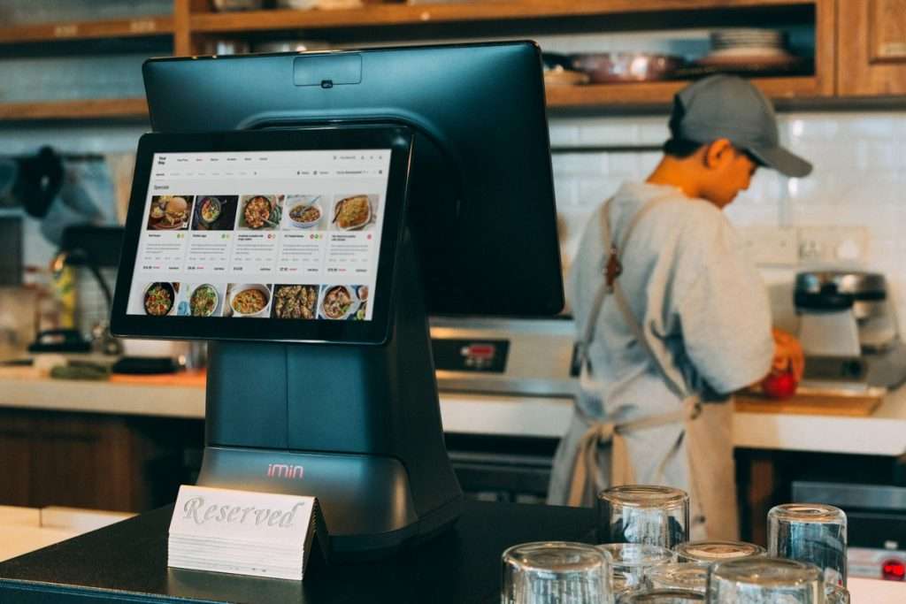 Ecrã tátil de pedidos num restaurante, com um funcionário na cozinha