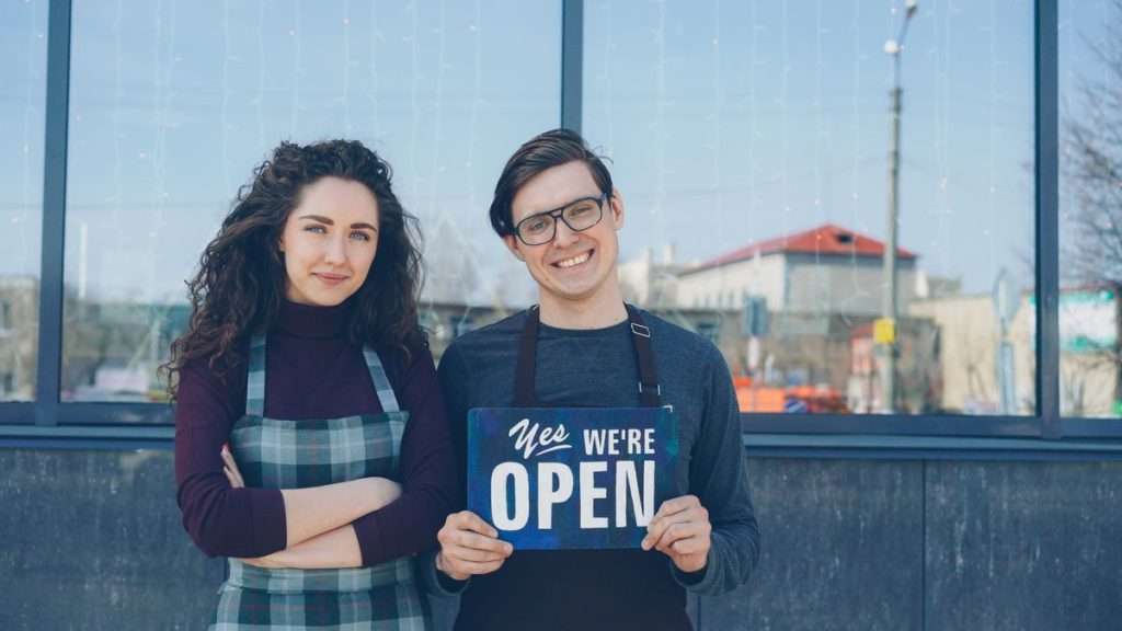 Dois comerciantes de avental a segurar um letreiro de loja aberta